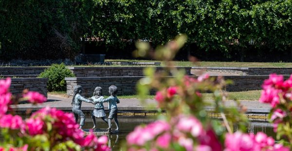 Bronze sculpture of children holding hands in a water feature in Rathfarnham Village Park, framed by flowers and seating